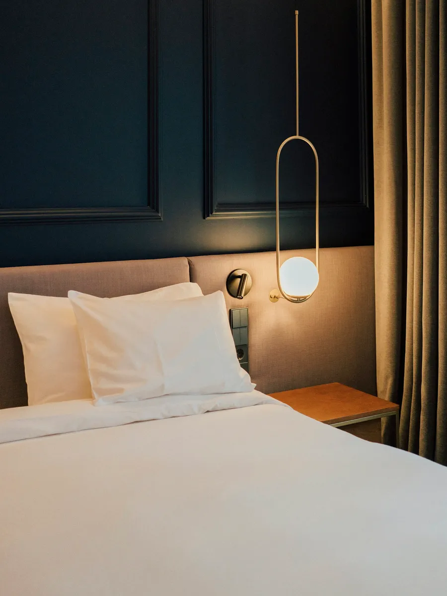 Hotel bed with white linen and a brass lamp against a dark wall
